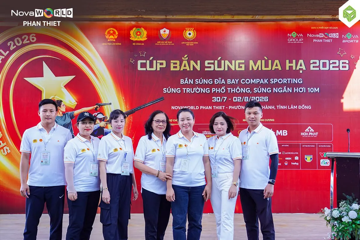 Nhóm chín người mặc áo trắng đứng chụp ảnh trước phông đỏ lớn của Cúp Bắn súng Mùa Hạ 2026